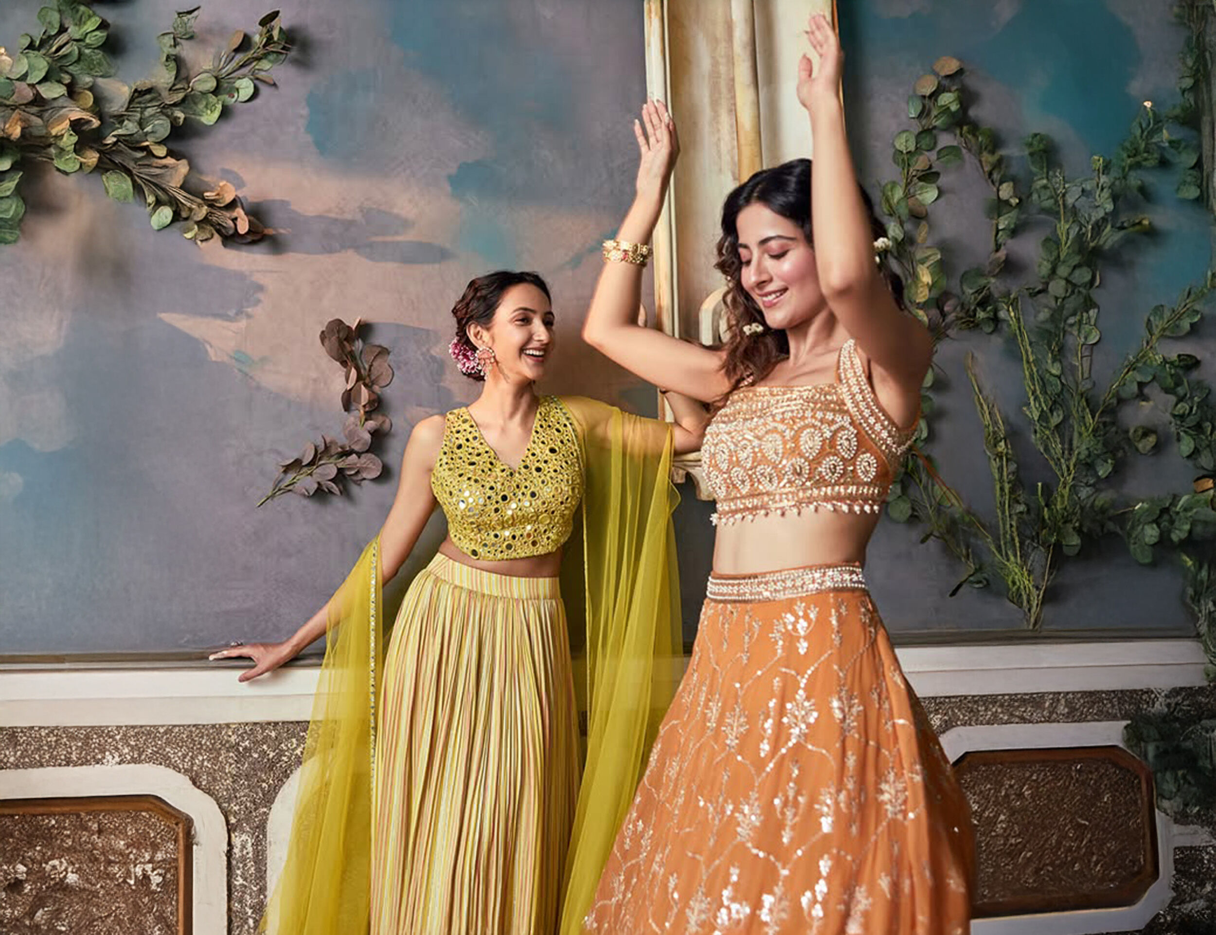 Two women celebrating in yellow and orange embroidered lehengas, showcasing modern festive wear fabrics.